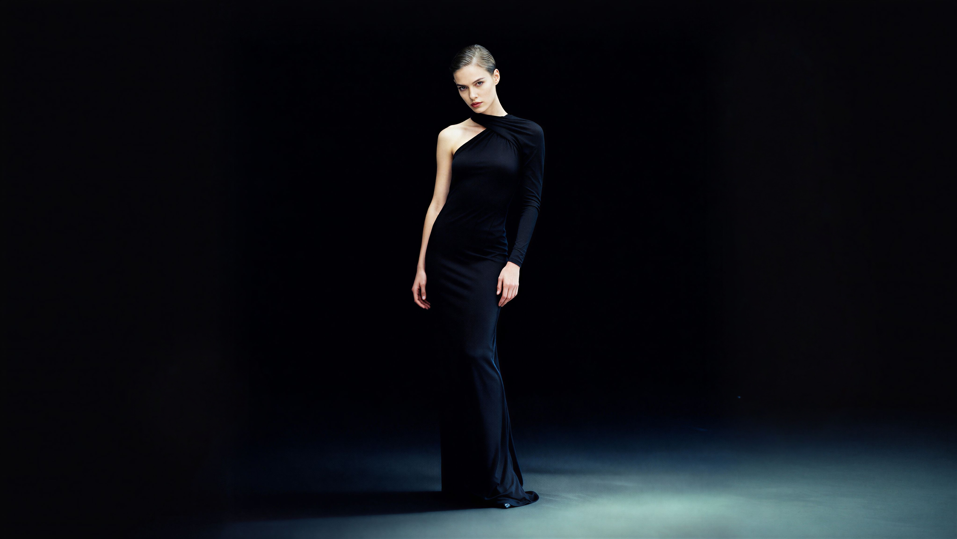 Woman in a black one-shoulder designer dress standing against a dark background